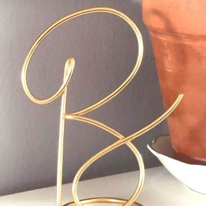 May include: A gold-coloured metal letter "B" sculpture. The letter is crafted from a continuous wire, forming a cursive design. The sculpture stands on a white surface, with a grey background and a terracotta pot with a plant visible in the background.