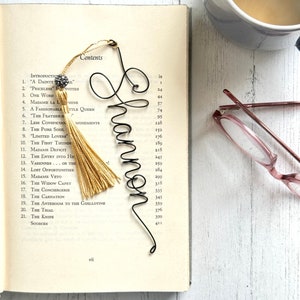 May include: A black wire bookmark shaped like the name "Shannon" with a gold tassel. The bookmark is placed on an open book with a table of contents listing chapter titles.