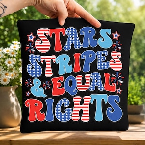以下が含まれることがあります： 黒い正方形のクッションで、太字のレトロなフォントで「STARS STRIPES & EQUAL RIGHTS」という文字がプリントされています。文字は赤と白の横縞、白星の入った青、単色の赤や青で装飾されており、小さな星のアイコンもあしらわれています。