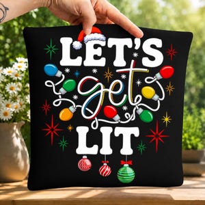 May include: A black square throw pillow cover featuring the white text LET'S get LIT with a Santa hat on the letter T. The design includes colorful Christmas light bulbs, red and green starbursts, and three hanging striped Christmas ornaments.