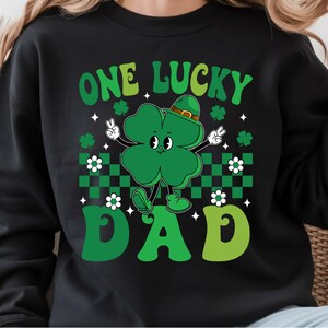 May include: Black sweatshirt with a St. Patrick's Day design. The graphic features a green four-leaf clover character wearing a hat and making a peace sign. The text reads "ONE LUCKY DAD" in green.