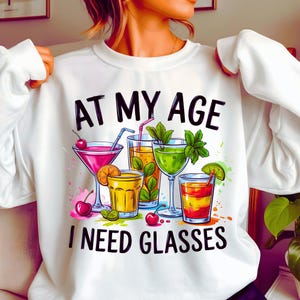 May include: White sweatshirt with the text "AT MY AGE I NEED GLASSES" above a colorful illustration of cocktails. The graphic includes a martini, a highball, and other mixed drinks with cherries, mint, and citrus garnishes.
