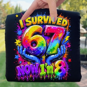 Könnte beinhalten: Schwarzes T-Shirt mit einem farbenfrohen Aufdruck "I Survived 67 Now I'm 8". Die Zahlen 67 sind in Regenbogenfarben gehalten und werden von blauen Händen gehalten. Das Design enthält Sterne und Blitze.