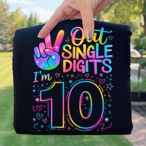 May include: Black t-shirt with a colorful peace sign and the text "Out Single Digits I'm 10". The design features a rainbow color scheme with hearts and stars. The shirt is held up by a hand.