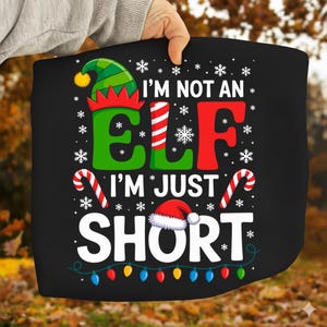 Pode incluir: Gráfico preto com o texto "I'M NOT AN ELF I'M JUST SHORT" em branco, verde e vermelho. O design inclui um chapéu de elfo, flocos de neve, bengalas doces, um gorro de Papai Noel e luzes de Natal coloridas.
