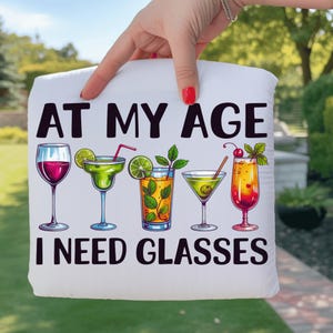 Puede incluir: Cojín blanco con el texto "AT MY AGE I NEED GLASSES" e ilustraciones de cócteles. Los cócteles incluyen vino tinto, margarita y otras bebidas mezcladas con adornos como lima y cerezas.