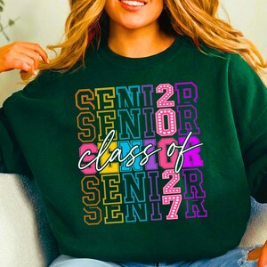 May include: Dark green sweatshirt featuring the text "SENIOR CLASS OF 2027" in a vibrant, stacked design. The text is in yellow, pink, purple, and blue, with a white cursive "class of". A fashionable item for graduation.
