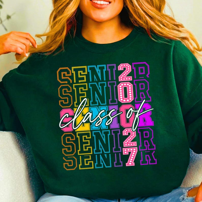 May include: Dark green sweatshirt featuring the text "SENIOR CLASS OF 2027" in a vibrant, stacked design. The text is in yellow, pink, purple, and blue, with a white cursive "class of". A fashionable item for graduation.