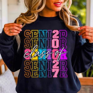 May include: Black sweatshirt featuring the text "SENIOR 2023" in a vibrant, layered design. The words "class of" are written in a cursive font across the centre. The sweatshirt is a casual style.