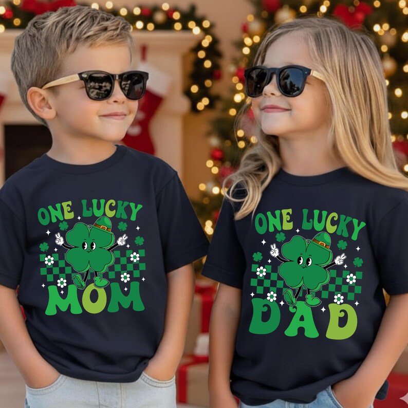 May include: Two children wearing navy blue t-shirts with a St. Patrick's Day design. The shirts feature a green four-leaf clover character with the text "ONE LUCKY MOM" and "ONE LUCKY DAD" in green. The clover has a hat and is surrounded by white flowers.