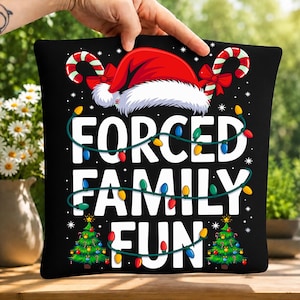 May include: A black square pillow featuring the white text FORCED FAMILY FUN. The design includes a red Santa hat, two candy canes with red bows, and two green Christmas trees. A string of colorful Christmas lights wraps around the text.