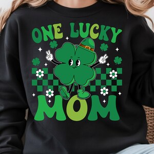 May include: Black sweatshirt with a green graphic of a four-leaf clover wearing a hat and giving a peace sign. The text "ONE LUCKY MOM" is in green. White flowers and green and black checkered pattern are also present.
