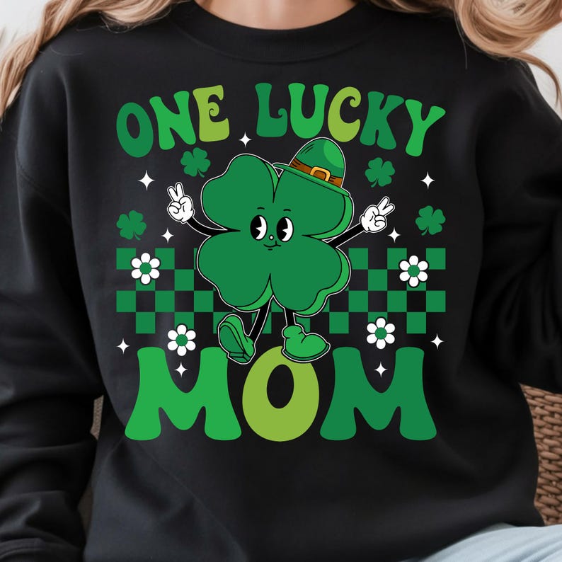 May include: Black sweatshirt with a green graphic of a four-leaf clover wearing a hat and giving a peace sign. The text "ONE LUCKY MOM" is in green. White flowers and green and black checkered pattern are also present.