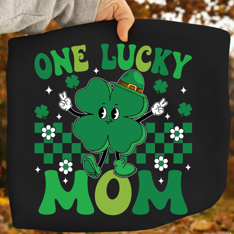 May include: Black graphic tee featuring a cartoon clover with a hat, giving peace signs. The text "ONE LUCKY MOM" is in green, with shamrocks and white flowers. The design has a retro, vintage style.