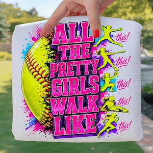 以下が含まれることがあります： ピンクと黄色の「ALL THE PRETTY GIRLS WALK LIKE THIS!」という文字が入った白いソフトボール枕。左側には黄色のソフトボールのグラフィックがあります。枕は手で持たれています。