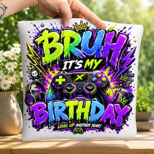May include: A square white throw pillow featuring a vibrant graffiti-style graphic with the text "BRUH IT'S MY BIRTHDAY" and "LEVEL UP ANOTHER YEAR!" in neon green, blue, and purple. The design includes a black game controller, headphones, and crowns.