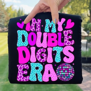 May include: Black t-shirt with the text "IN MY DOUBLE DIGITS ERA" in a retro font. The text features a mix of pink, teal, and leopard print patterns. A disco ball graphic is at the bottom.