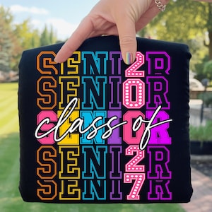 May include: Black t-shirt featuring the text "SENIOR" and "class of 2027" in a vibrant, multi-coloured design. The lettering includes orange, yellow, pink, blue, purple, and white. The shirt is held up by a hand.