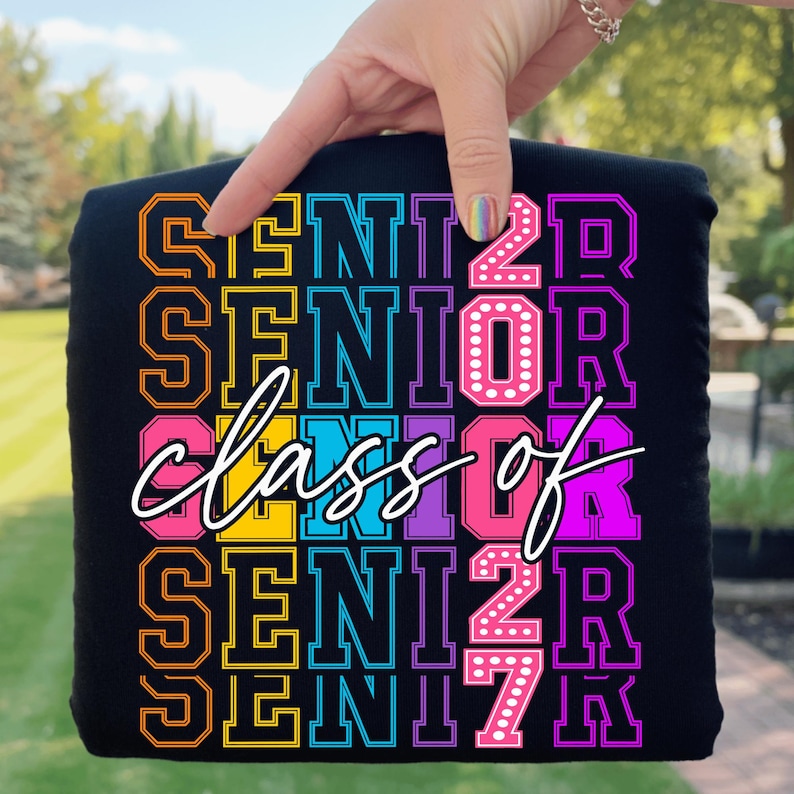 May include: Black t-shirt featuring the text "SENIOR" and "class of 2027" in a vibrant, multi-coloured design. The lettering includes orange, yellow, pink, blue, purple, and white. The shirt is held up by a hand.