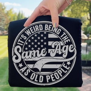 May include: Black t-shirt with a white graphic design. The design features a circular emblem with the text "IT'S WEIRD BEING THE SAME AGE AS OLD PEOPLE" surrounding an American flag design.