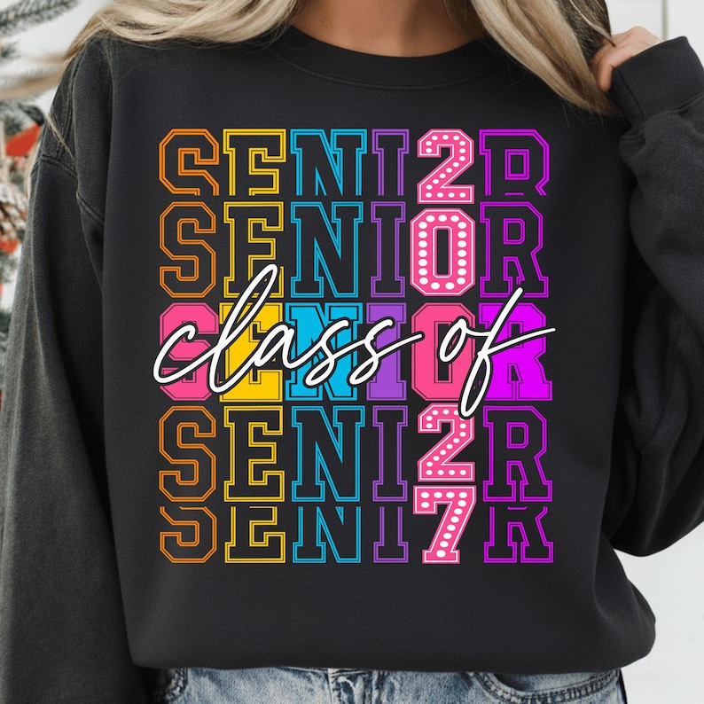 May include: Black sweatshirt featuring the text "SENIOR class of 2027" in a vibrant, layered design. The text is in shades of orange, yellow, pink, purple, and blue, with a white cursive "class of".