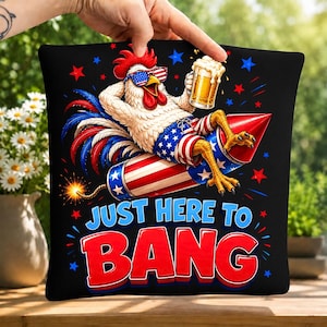 May include: A black square pillow featuring a white rooster wearing American flag sunglasses, blue and white star-patterned shorts, and holding a beer mug while riding a red, white, and blue firework. Text reads JUST HERE TO BANG.