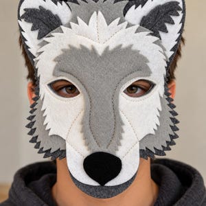 May include: A felt wolf mask featuring layered gray and white fabric sections, pointed ears, and a solid black nose. The mask is constructed with visible stitching along the edges of the felt pieces and includes two eye openings for visibility.