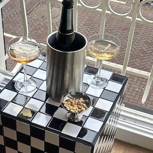 Puede incluir: Una mesa auxiliar de azulejos negros y blancos con una botella de champán en una nevera de plata, dos copas de champán y un pequeño cuenco de frutos secos.