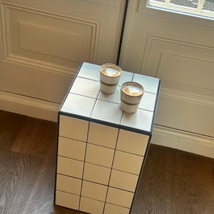 Può includere: Tavolino laterale piastrellato, con cornice nera e superficie piastrellata bianca. Due tazze di caffè con latte art sopra. Il tavolo è angolato, con una finestra e un pavimento in legno sullo sfondo.