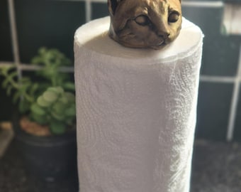 May include: A decorative paper towel holder featuring a cat head sculpture in a bronze finish. The cat head sits atop a roll of white paper towels, with a gold-colored base. The holder adds a unique touch to any kitchen.