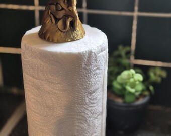May include: A decorative paper towel holder featuring a detailed, golden-colored rooster head. The rooster sits atop a roll of white paper towels. The holder adds a rustic touch to any kitchen.