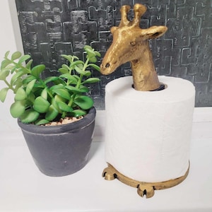 May include: Gold giraffe-shaped toilet paper holder with a white roll of toilet paper. The holder has a round base with three feet.