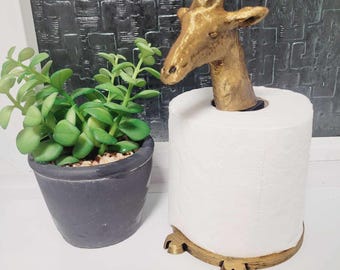 May include: Gold giraffe-shaped toilet paper holder with a white roll of toilet paper. The holder has a round base with three feet.