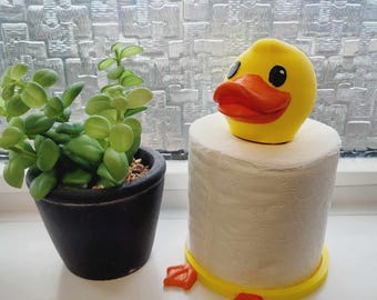 May include: Yellow rubber duck toilet paper holder with orange feet. The holder is on a white window sill with a potted plant in the background.
