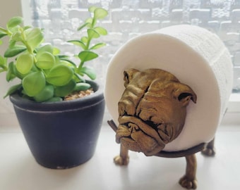 May include: A gold bulldog figurine holding a roll of white toilet paper. The bulldog is standing on a black metal stand.