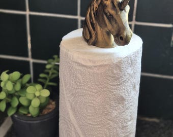 May include: A white paper towel roll with a gold-colored base and a decorative unicorn head topper. The unicorn has a horn and detailed mane. A small potted plant is in the background.