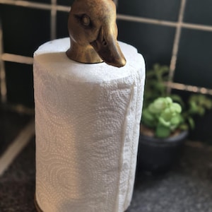 May include: A decorative paper towel holder featuring a duck head design in a bronze finish. The holder is topped with a duck head and holds a roll of white paper towels. The holder is on a countertop.