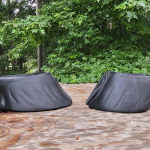 May include: Two black, 3D-printed horse hoof boots are displayed side-by-side on a wooden surface. The boots are designed to protect a horse's hooves. The background features lush green foliage.