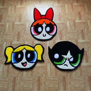May include: Three cartoon character rugs featuring the Powerpuff Girls: Blossom, Bubbles, and Buttercup. The rugs are made of a soft, plush material and feature the characters' signature colours and facial expressions.