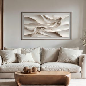 May include: A framed abstract art piece with a neutral color palette hangs above a cream-colored sofa. The artwork features flowing, layered forms in shades of white and beige. The living room setting includes a wooden coffee table and decorative elements.