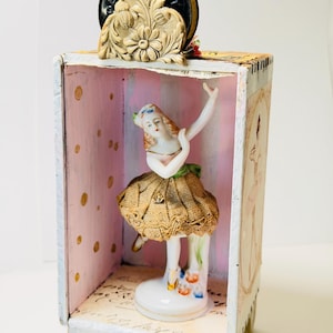 May include: A vintage-style shadow box featuring a porcelain ballerina figurine with a lace skirt. The box has a pink interior with gold polka dots and a decorative top piece. The box is made of wood and paper.
