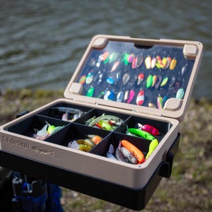 Può includere: Una scatola da pesca aperta, piena di esche colorate. La scatola beige e nera, con coperchio trasparente, mostra il testo "SJ design". Le esche sono di varie forme e colori.