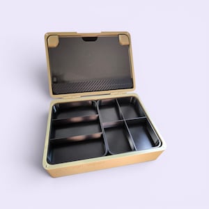 May include: An open, gold-colored organizer box with a black interior. The box is divided into multiple compartments of varying sizes. The lid is open, revealing a dark interior with a textured pattern. The box is designed for storage and organization.