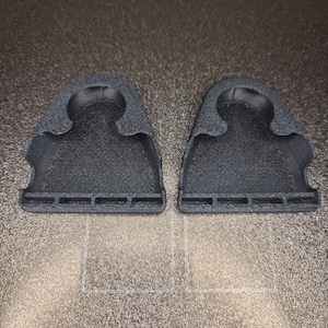 May include: Two black, textured plastic objects, each with a curved top and a series of rectangular cutouts along the bottom edge. The objects are positioned side-by-side on a textured, brown surface.