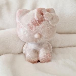 Puede incluir: Una figurita de Hello Kitty de piedra blanca y rosa. La figurita está sentada sobre una superficie blanca y esponjosa.