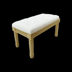 Peut inclure: Un petit banc rectangulaire avec une assise rembourrée blanche et un cadre en bois beige clair. Les pieds sont cylindriques et le cadre présente des moulures décoratives. Le banc est posé sur un fond noir.