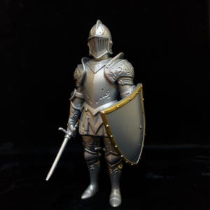 Puede incluir: Figura de caballero plateado, con casco, armadura, escudo y espada. El escudo tiene un borde dorado. El caballero está en una postura de combate. La figura es una representación detallada de una armadura medieval.