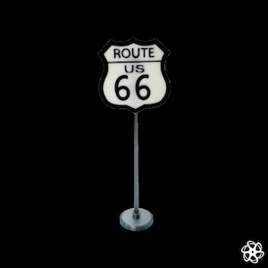 Puede incluir: Un letrero en miniatura de la Ruta 66 con fondo blanco y borde negro. El letrero dice "ROUTE US 66" en texto negro. Está montado sobre un soporte de metal plateado con una base redonda. Un símbolo clásico de los viajes por carretera americanos.
