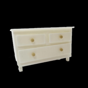 Op de afbeelding: Een kleine, crèmekleurige miniatuur ladekast met vier lades en goudkleurige knoppen. De kast heeft een eenvoudig ontwerp met een rechthoekige vorm en vier korte poten. De achtergrond is effen zwart.