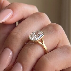 Könnte beinhalten: Ein Goldring mit einem einzelnen rechteckigen, im Kissenschliff geschliffenen, klaren Edelstein in einer Zargenfassung. Die Ringschiene ist schmal und weist ein geteiltes Design auf, bei dem sich das Metall in zwei Arme teilt, bevor es den Stein erreicht.